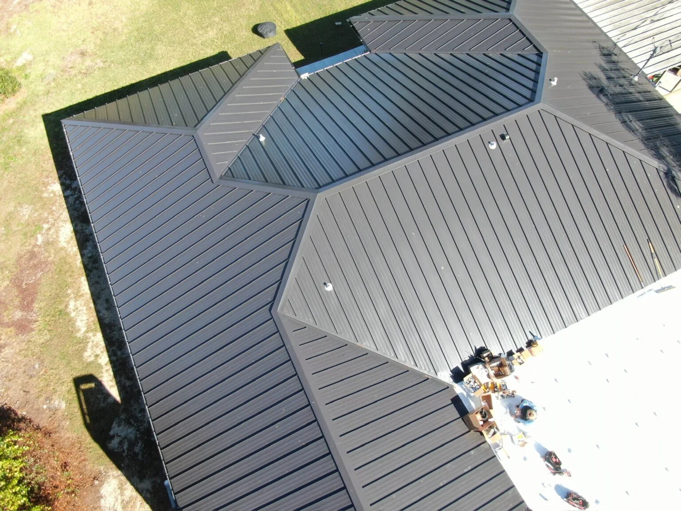 Greentek project photo — a drone view looking down on the valleys and hips of a dark standing-seam metal roof, with the crew and materials staged at the edge