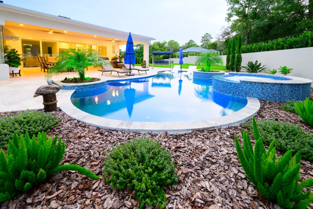 Nic’s Pool Construction project photo — a curved freeform pool with a raised blue mosaic spa, travertine deck and planted bark beds at dusk