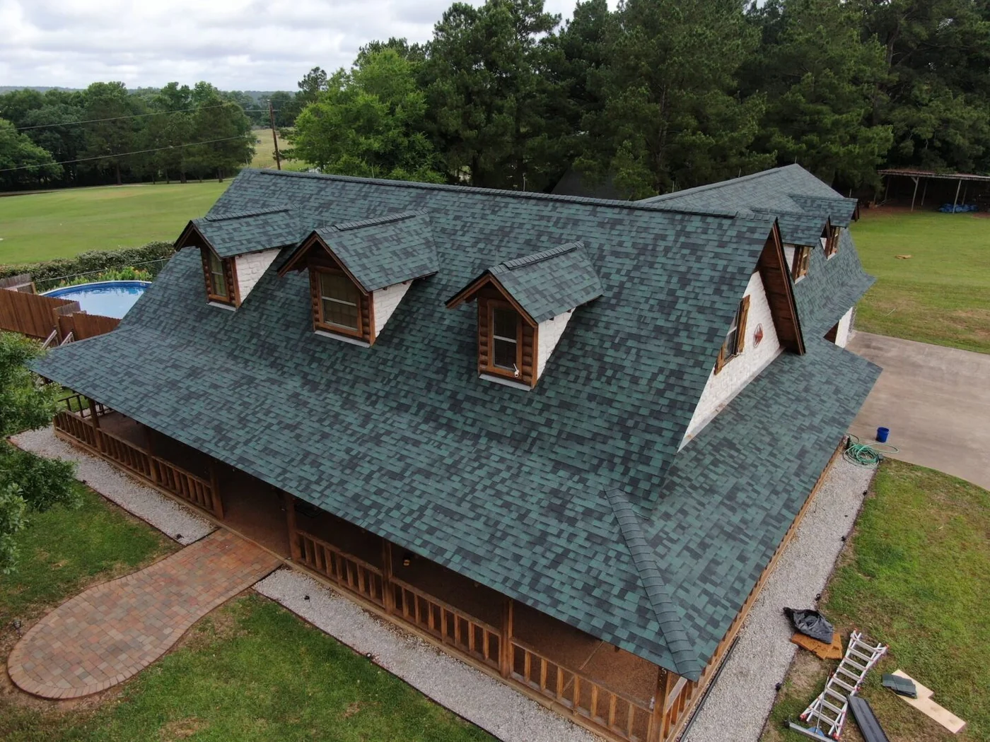 Roof Ryders project photo — a drone view of a dormered ranch house in Lufkin, Texas with a new green architectural shingle roof, a pool and deck below